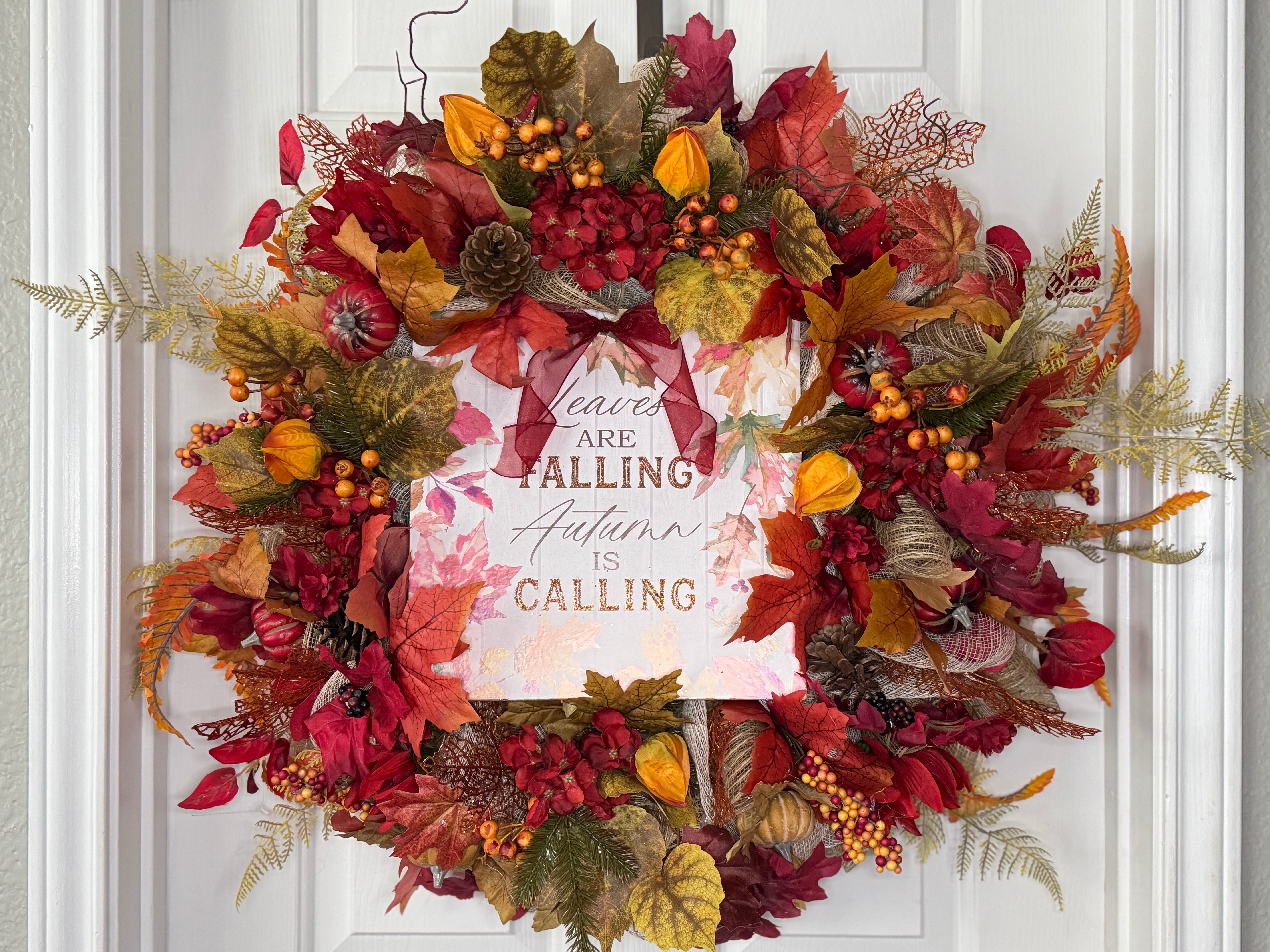 Rustic burgundy and pumkin colored fall leaves, flowers, and greenery adorn a round mesh wreath with a central autumn/Fall inspired sign, Burgundy matching ribbon