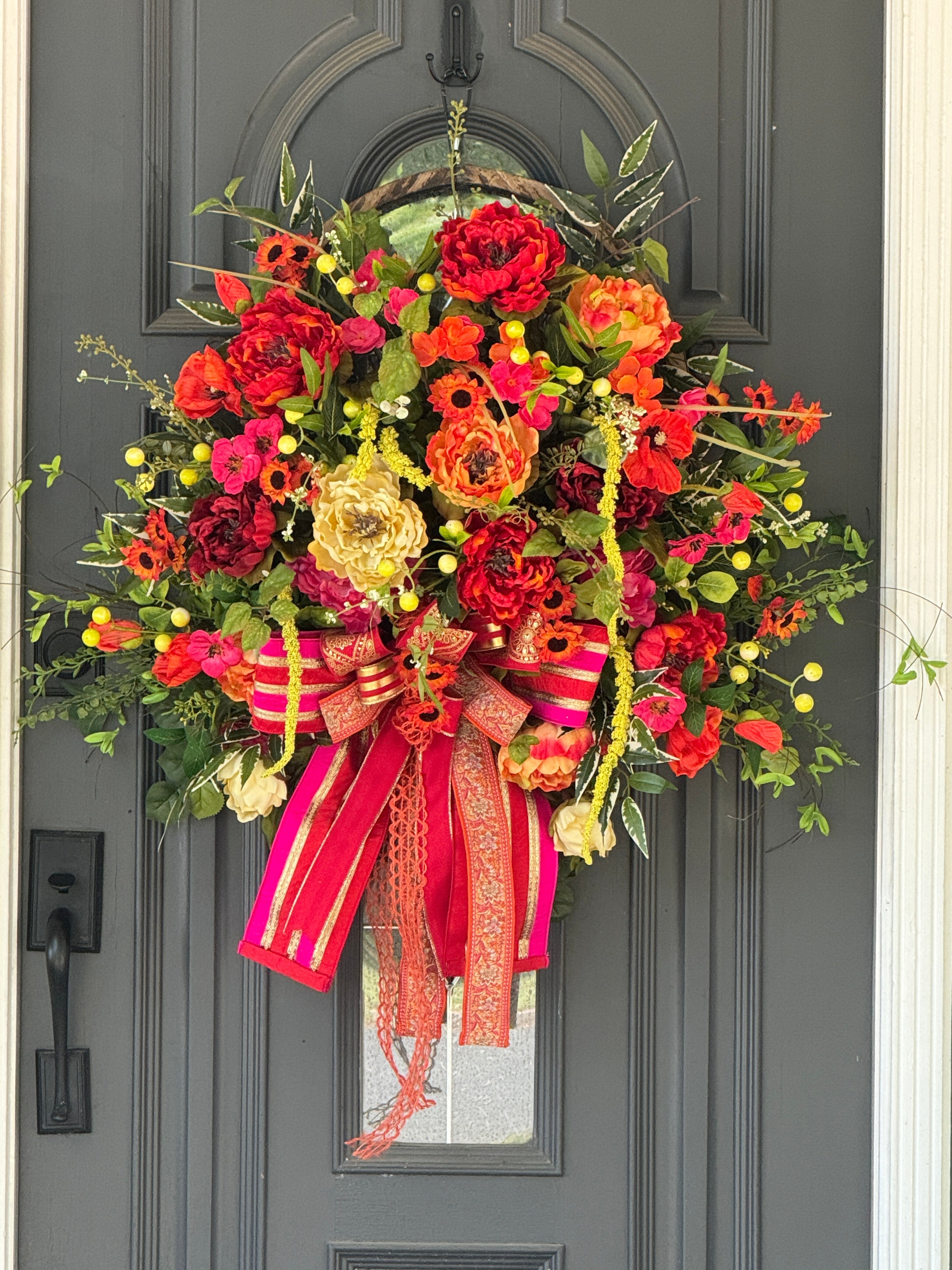 Large oval wreath with bright red , fuchsia, and orange flowers, greenery and beutifully matched bow with a cascade of ribbon